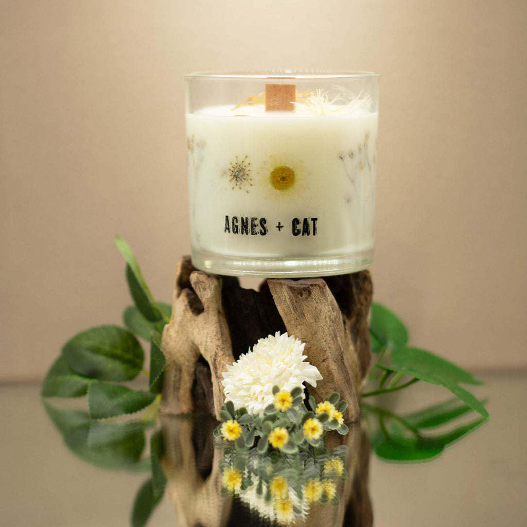 Candle in a glass jar with 'AGNES + CAT' branding on a wooden stand with flowers and leaves.