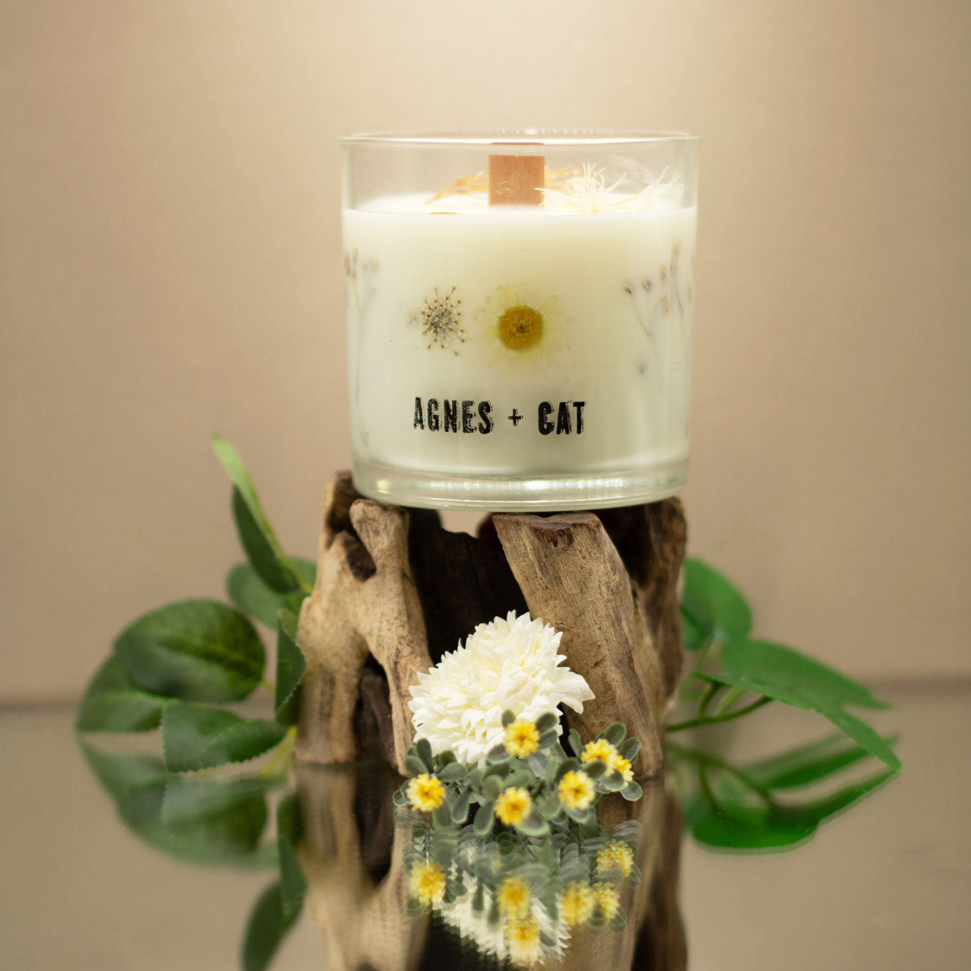 Candle in a glass jar with 'AGNES + CAT' branding on a wooden stand with flowers and leaves.